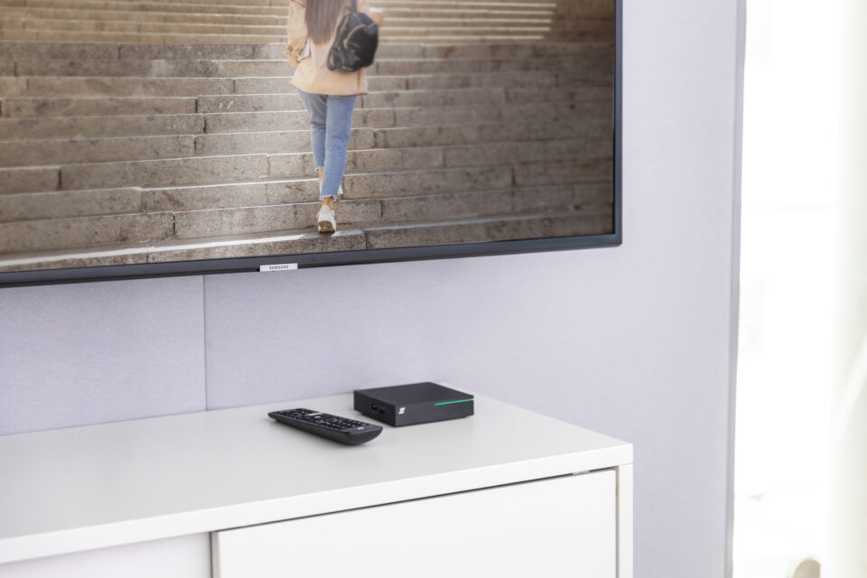 Auf einem weißen Sideboard liegen eine Fernbedieung und ein Receiver in schwarz. Das TV-Gerät an der Wand zeigt ein Programm.