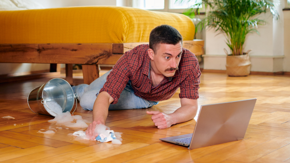 Junger Mann putzt den Boden und schaut dabei TV auf seinem Laptop