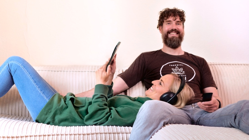 Paar, bestehend aus einem Mann und einer Frau, entspannt gemeinsam auf dem Sofa. Die Frau trägt Kopfhörer und schaut auf ihr Tablet, der Mann schaut TV