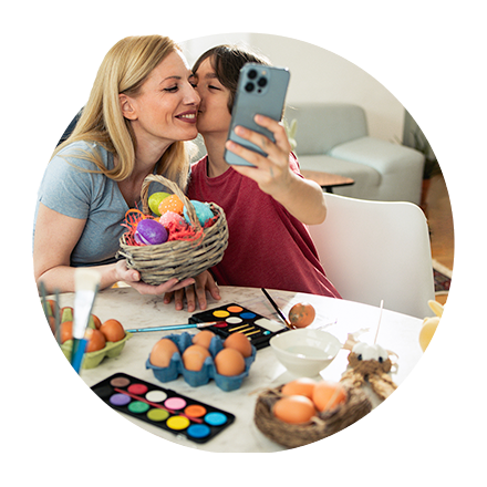 Das Bild zeigt zwei Personen, die an einem Tisch sitzen. Eine Person hält ein Smartphone und macht ein Bild von beiden Personen. Auf dem Tisch befinden sich mehrere Eier in einer blauen Eierschachtel sowie einige lose Eier. Es gibt auch eine Palette mit Farben und einen Pinsel, was darauf hindeutet, dass die Personen möglicherweise Eier bemalen.