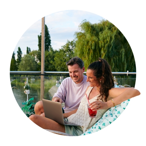 Junges Paar sitzt auf Dachterrasse vor dem Laptop. Im Hintergrund ist der Bodensee zu sehen. Die Sonne scheint und die Frau trinkt einen roten Cocktail.