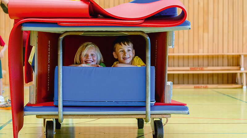 Zwei Kinder sitzen lachend in einer selbstgebauten Höhle aus Turnmatten und Geräten in einer Sporthalle.