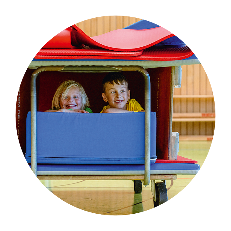 Zwei Kinder sitzen lachend in einer selbstgebauten Höhle aus Turnmatten und Geräten in einer Sporthalle.