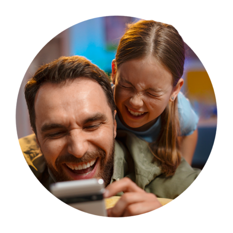 Ein lachender Mann liegt auf dem Sofa und schaut mit einem Kind gemeinsam auf ein Smartphone. Beide wirken fröhlich und entspannt.